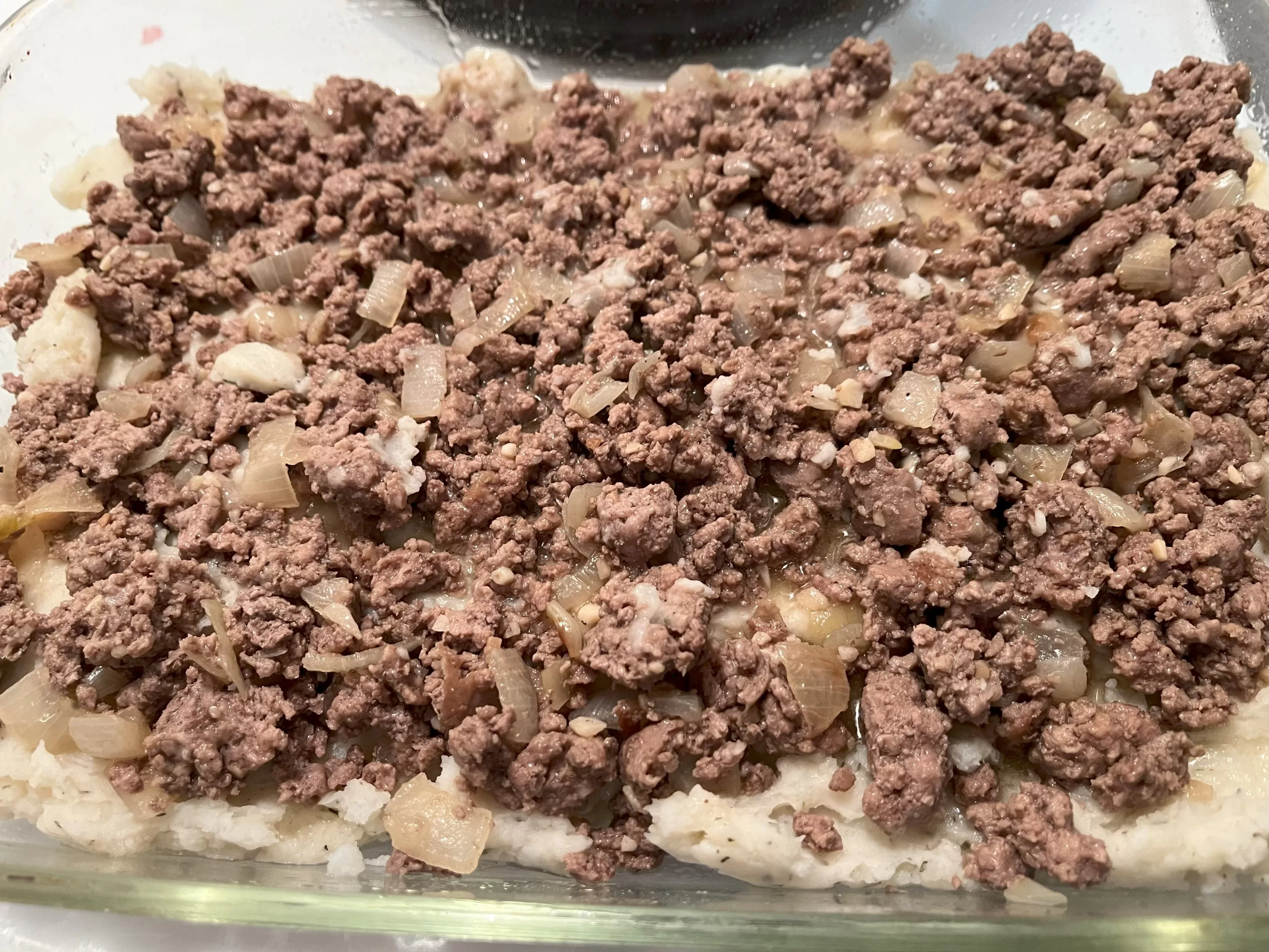 Ground beef, onions, and garlic on top of mashed potatoes in a glass dish.