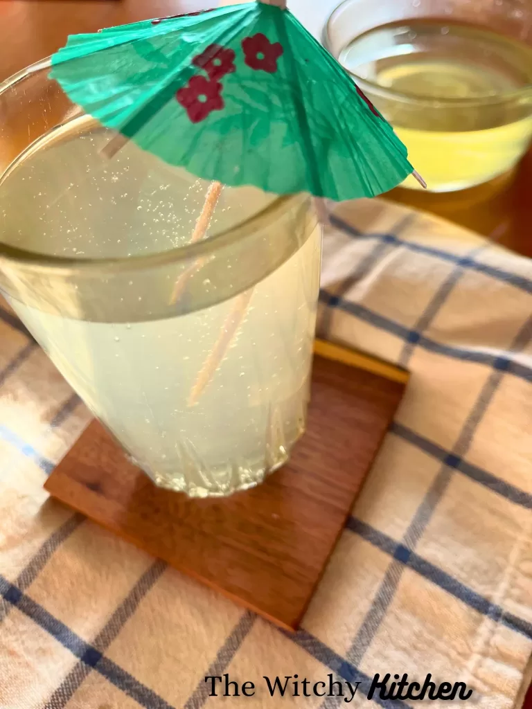 A tall glass of homemade ginger ale with a green paper umbrella inside and on the edge. The glass is on a wooden coaster, with a white towel that has blue lines making squares. A glass of yellow ginger syrup is in the background on a wooden table.