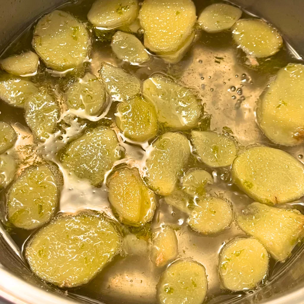 A mixture of ginger slices, lime zest and juice, water, and sugar boiling in a metal pot.
