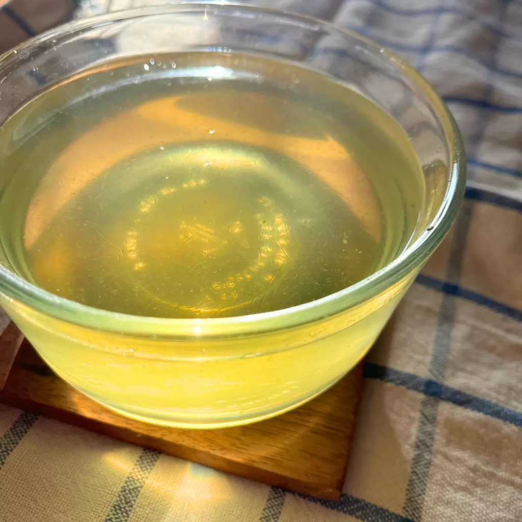 A round clear glass dish filled with yellow ginger syrup. The dis is on a square-shaped wooden coaster, on top of a white towel with blue lines that make a square/checker design.