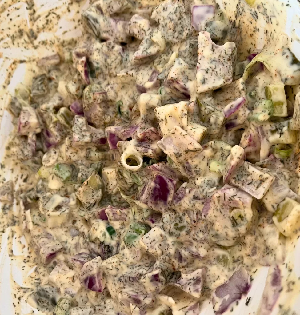 Red onions, green onions, pickles, mayonnaise, Dijon mustard, apple cider vinegar, dill, and other seasonings mixed together in a white bowl.