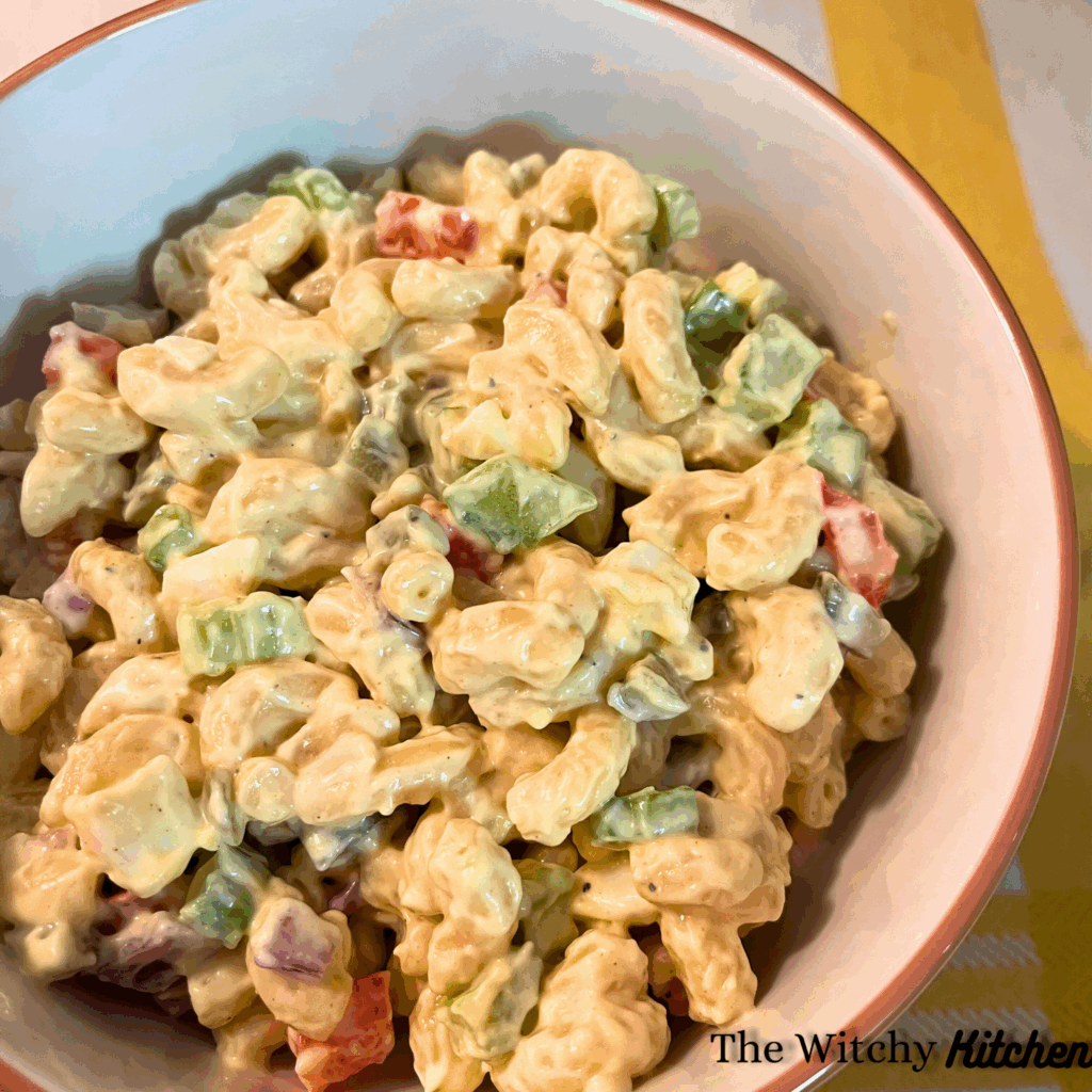 Macaroni salad with macaroni, vegetables (celery, red bell pepper, red onion, sweet gherkins), hard-boiled eggs and a yellow-white dressing, in a white bowl on top of a yellow and white cloth.