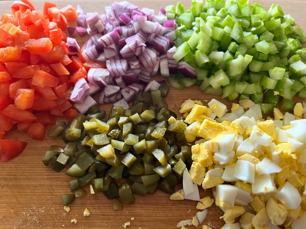 Piles of diced red bell pepper, red onions, celery, sweet gherkins, and hard-boiled eggs on a wooden cutting board.