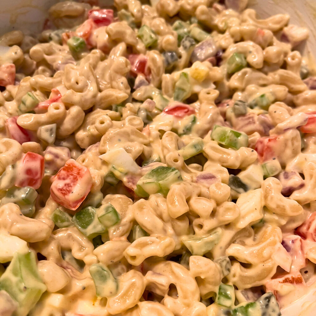 A close up of macaroni salad [with macaroni, vegetables (celery, red bell pepper, red onion, sweet gherkins), hard-boiled eggs and a yellow-white dressing].