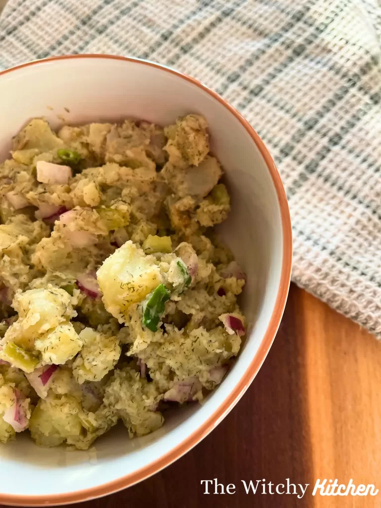 Potato salad in a white bowl on a wooden board with a checkered tea towel on the board.