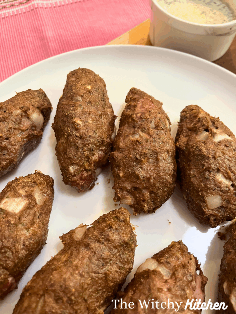 Chicken kofta kebabs on a white plate. A white bowl with garlic-yogurt sauce is in the top right corner and a pink placemat is in the top left corner.