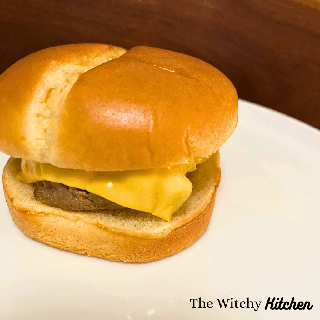 A hamburger patty between 2 buns with a slice of cheese, on a white plate with a brown background.