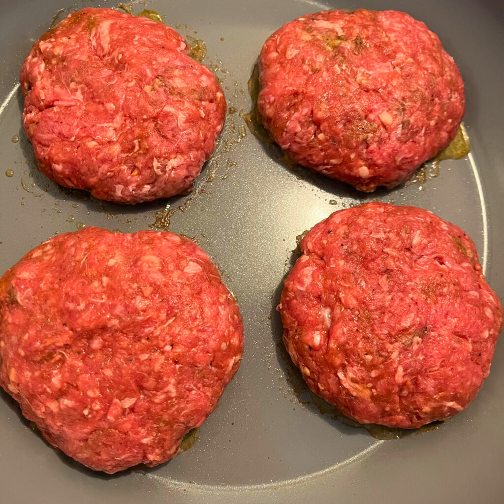 4 raw hamburger patties cooking in a grey frying pan.