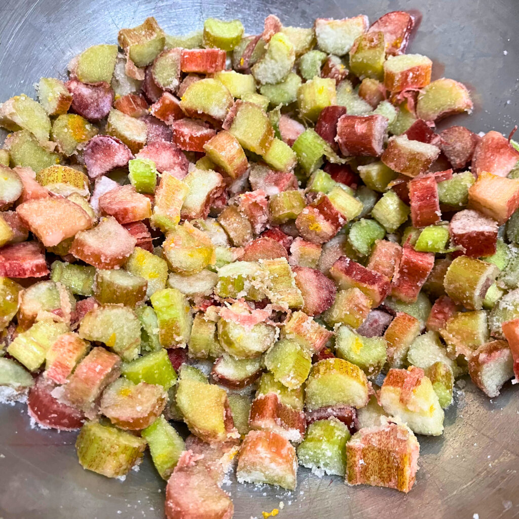 Pieces of rhubarb covered in corn starch, sugar, salt and orange zest in a metal mixing bowl.