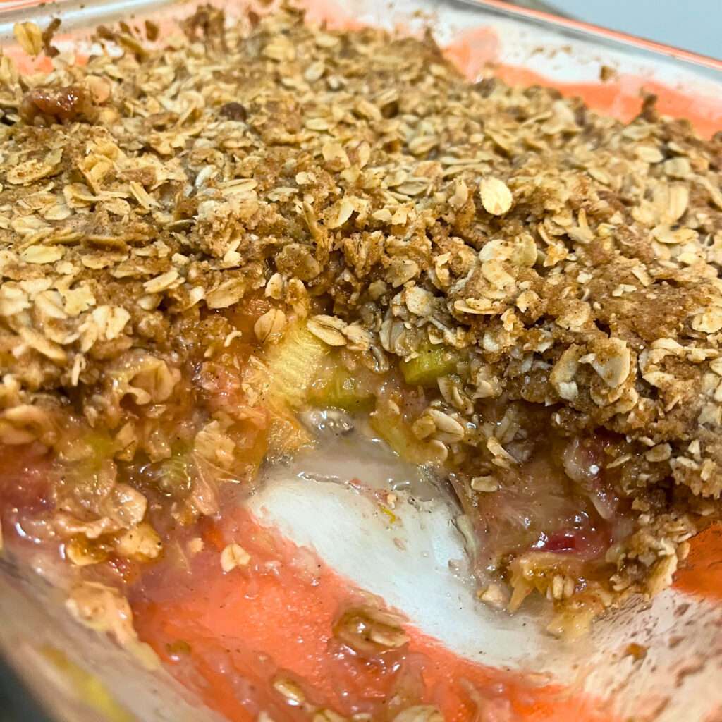 Rhubarb crisp with pieces of rhubarb and juices, topped with oats and brown pieces, in a clear glass baking dish on an orange and white striped towel.